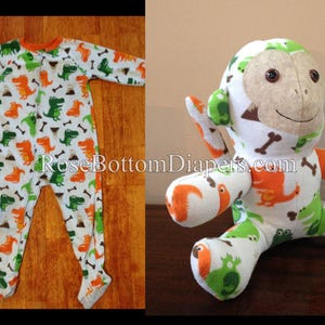 Op de afbeelding: Een witte, oranje en groene dinosaurus-geïnspireerde voetpyjama en een bijpassende knuffel. De pyjama en aap hebben een herhalend patroon van oranje en groene dinosaurussen en botvormen. De tekst "RoseBottomDiapers.com" is zichtbaar.