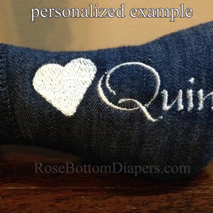 May include: Close-up of a denim fabric item with white embroidered details. A white heart is embroidered next to the name "Quinn" in white script. The text "personalized example" is at the top.