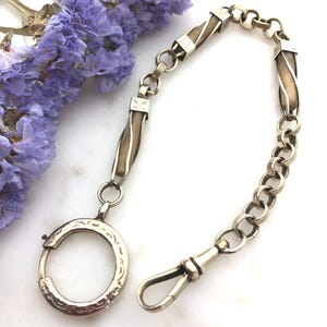 May include: A silver-toned watch chain featuring a circular engraved pendant, a series of metallic links, and two elongated, twisted beige segments. The chain terminates in a small, secure hook clasp, providing a functional and decorative accessory.