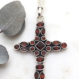 Puede incluir: Un collar con un colgante de cruz de plata adornado con un diseño de piedras de granate rojo. La cruz está decorada con varias piedras de granate de forma ovalada.