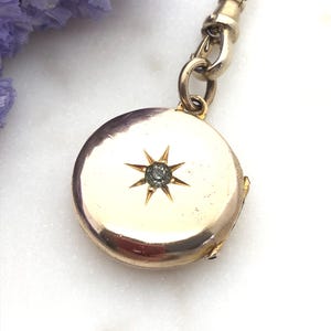 Könnte beinhalten: Ein goldfarbener, runder Medaillon-Anhänger mit einem Sternmuster und einem zentralen Edelstein. Das Medaillon ist an einer zarten Goldkette und einem Verschluss befestigt. Der Hintergrund umfasst lila Blumen und eine weiße Marmoroberfläche.