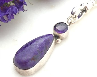 May include: A silver pendant featuring a large, teardrop-shaped purple stone and a smaller, round purple stone. The pendant is connected to a silver chain. Purple flowers are in the background.