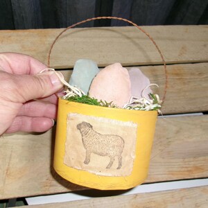 May include: A yellow, handcrafted container with a sheep illustration on the front. The container holds decorative eggs and faux greenery. A brown wire handle arches over the top. The container is held by a hand.