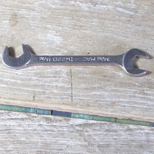 May include: A silver MAC wrench with two different sized open ends. The wrench is lying on a weathered wooden surface. The text "MAC" and "1W22D" are visible on the wrench.