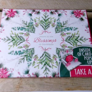 May include: A decorative Christmas card box with a white background and a pattern of green leaves, pink flowers, and red accents. The text "Share in the Blessings of Christmas" is displayed. A smaller image shows a pop-up card.