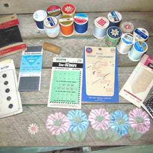 May include: A collection of vintage sewing supplies, including spools of thread in various colors, buttons, seam binding, snaps, and needle assortments. The items are arranged on a wooden surface, with floral embroidery and trim.