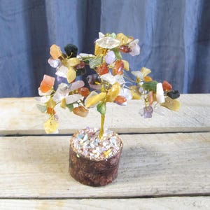 May include: A decorative gemstone tree with a gold-colored wire trunk and branches. The tree's leaves are made of various colorful gemstones, including orange, yellow, white, and green. The tree sits in a small, brown, cylindrical pot filled with small stones.