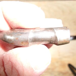 May include: A metallic bullet with a pointed tip and a cylindrical body. The bullet has a tarnished, aged appearance with visible rust-colored spots. The bullet is held in a person's hand.