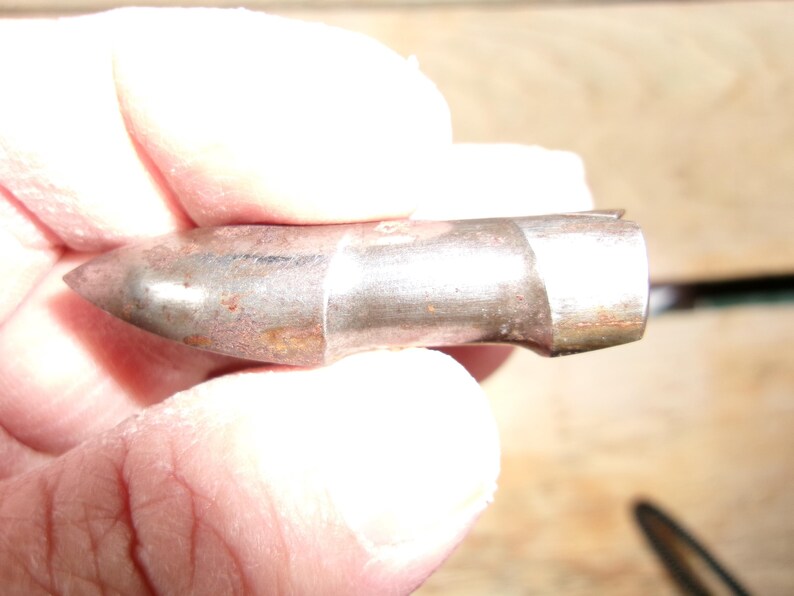 May include: A metallic bullet with a pointed tip and a cylindrical body. The bullet has a tarnished, aged appearance with visible rust-colored spots. The bullet is held in a person's hand.