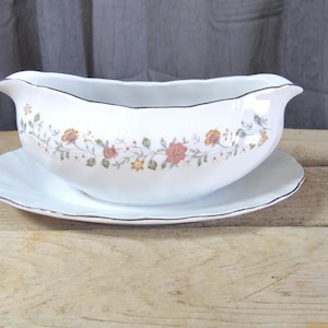 May include: White porcelain gravy boat with a matching saucer. The boat features a floral design in shades of orange, pink, and green, with a black trim. The set is placed on a wooden surface.