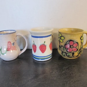 May include: Three ceramic mugs with different designs. The first mug is white with a pink and green floral design. The second mug is white with three red strawberries. The third mug is yellow with a black and red floral design.