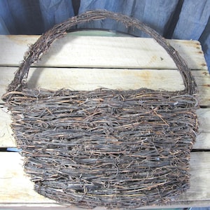 May include: A rustic, brown woven basket with a curved handle. The basket is made of intertwined twigs and branches, creating a textured surface. The handle is also made of woven twigs. The basket sits on a wooden surface.