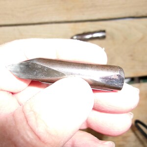 May include: A metal tool with a sharpened, angled tip. The tool has a cylindrical body with a dark, metallic finish. The tip appears to be made of a different, lighter-colored metal. The tool is held in a hand, with a second tool visible in the background.