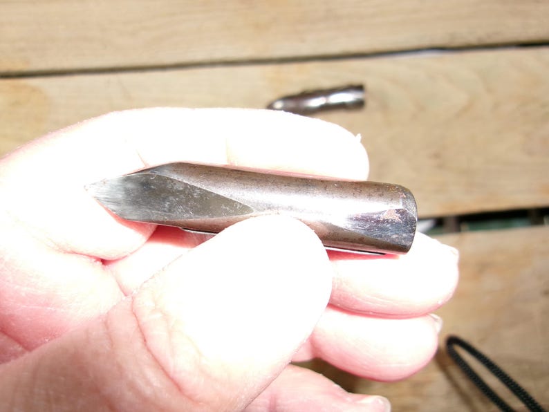 May include: A metal tool with a sharpened, angled tip. The tool has a cylindrical body with a dark, metallic finish. The tip appears to be made of a different, lighter-colored metal. The tool is held in a hand, with a second tool visible in the background.