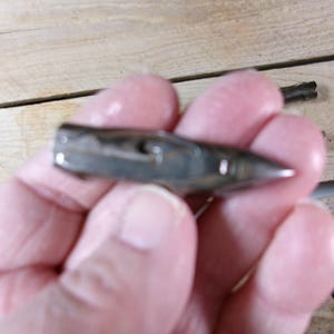 May include: A small, silver-colored bullet-shaped pendant with a pointed tip. The pendant is held in a person's hand, with a wooden surface in the background. A black cord is attached to the pendant.
