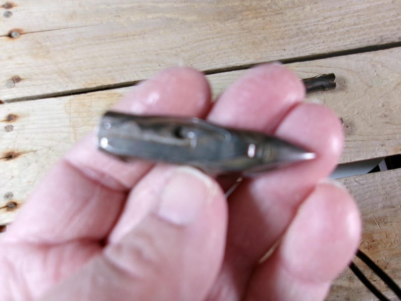 May include: A small, silver-colored bullet-shaped pendant with a pointed tip. The pendant is held in a person's hand, with a wooden surface in the background. A black cord is attached to the pendant.