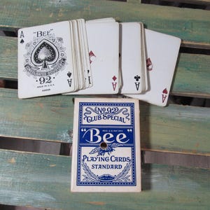 Puede incluir: Una baraja de cartas Bee con una caja azul y blanca, y varias cartas extendidas. Las cartas muestran el As de Picas y otros palos. La caja dice "No. 92 Club Special" y "Bee Playing Cards Standard."