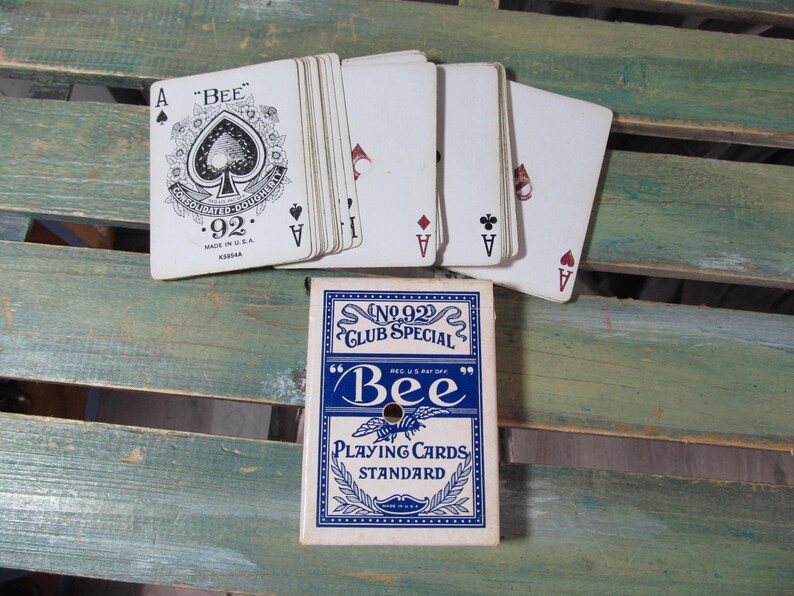 Puede incluir: Una baraja de cartas Bee con una caja azul y blanca, y varias cartas extendidas. Las cartas muestran el As de Picas y otros palos. La caja dice "No. 92 Club Special" y "Bee Playing Cards Standard."