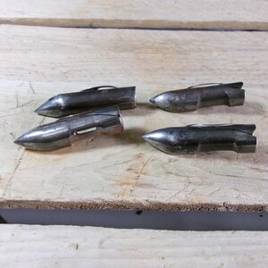 May include: Four vintage metal arrowheads with a silver finish are arranged on a light-colored wooden surface. Each arrowhead has a pointed tip and a small fin-like structure at the back. The arrowheads are likely for hunting or display.