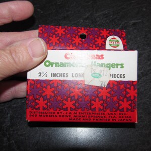 May include: A red and purple box with a snowflake pattern. The box contains 24 Christmas ornament hangers that are 2 1/2 inches long. The box is labeled "Christmas Ornament Hangers" and "Distributed by: J & M Enterprises (USA) Inc. 665 Mokena Drive, Miami Springs, Fla. 33166 Made and Printed in Japan."
