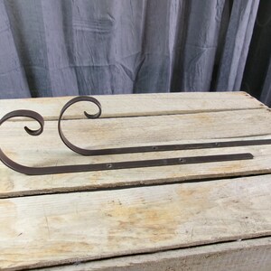 May include: Two dark brown metal shelf brackets featuring a flat horizontal arm with pre-drilled screw holes and a curved, scroll-shaped support arm that extends upward in a decorative loop design, providing structural reinforcement for shelving.