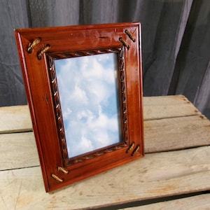 May include: A rectangular wooden picture frame with a reddish-brown finish. The frame has decorative rope accents at the corners and along the inner edge. The picture inside shows a blue sky with white clouds.