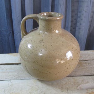 May include: A tan ceramic jug with a rounded body and a handle. The jug has a speckled surface and a glossy finish. It sits on a weathered wooden surface against a blue curtain backdrop. The jug is a decorative item.