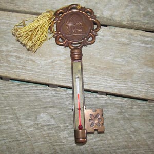 May include: A vintage key-shaped thermometer with a gold-colored metal body and a red and white thermometer scale. The key has a decorative top with a Native American head design and a gold tassel.