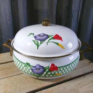 May include: White enamelware casserole dish with a lid, featuring purple and red floral designs. The dish has brass handles and a green lattice pattern around the base. A brass knob tops the lid.