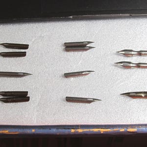 May include: A collection of nine calligraphy pen nibs in various shapes and sizes. The nibs are made of metal and are arranged on a light-colored surface. Some nibs have a split tip, while others have a rounded or pointed tip.