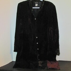 May include: A long, black velvet coat with a button-up front and long sleeves. The coat has a textured pattern and a contrasting trim around the collar and cuffs. The brand name "KOOS" is visible on the label.