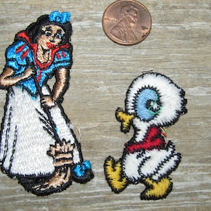 May include: Embroidered patches featuring Snow White in a blue and white dress and a cartoon duck in a red shirt. The patches are colorful and detailed, with black outlines. A penny is included for size reference.