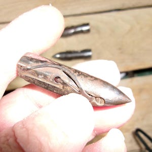 May include: A vintage, metallic bullet-shaped object with a pointed tip and a curved design. The object is held in a hand, with two similar objects visible in the background. The metal has a worn, aged appearance.