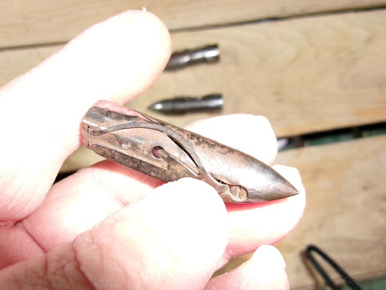 May include: A vintage, metallic bullet-shaped object with a pointed tip and a curved design. The object is held in a hand, with two similar objects visible in the background. The metal has a worn, aged appearance.