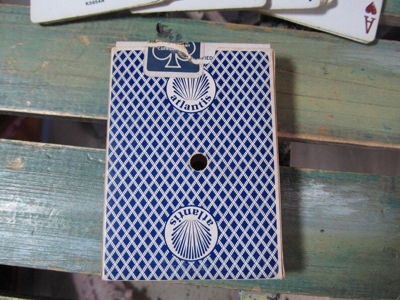 Puede incluir: Una baraja de cartas con un patr&oacute;n de diamantes azules y blancos en el reverso. La palabra "atlantis" est&aacute; impresa en el reverso de la baraja en un dise&ntilde;o circular. La baraja tiene un agujero en el centro.