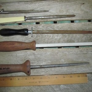 May include: A collection of vintage knife sharpeners and a wooden ruler. The sharpeners have wooden and black handles, and metal rods. One sharpener has a white handle. The wooden ruler is marked with inch measurements.