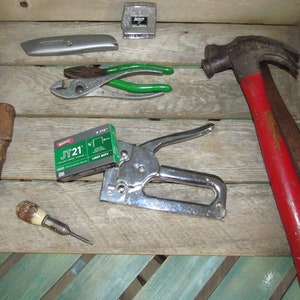 May include: A collection of tools including a hammer with a red handle, a staple gun, a box of staples, a pair of pliers, a utility knife, and two screwdrivers.