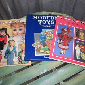 May include: Three books about toys and dolls. The book on the left is titled "Modern Collectibles Identifying & Value Guide". The book in the middle is titled "Modern Toys American Toys 1930-1980". The book on the right is titled "Precious Paper Dolls by Cynthia Perfect Bonner".
