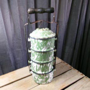 May include: A four-tiered stackable food container featuring a green and white marbled enamel finish. The cylindrical compartments are held together by a metal frame with a dark wooden handle at the top, allowing for easy transport of stacked meals.