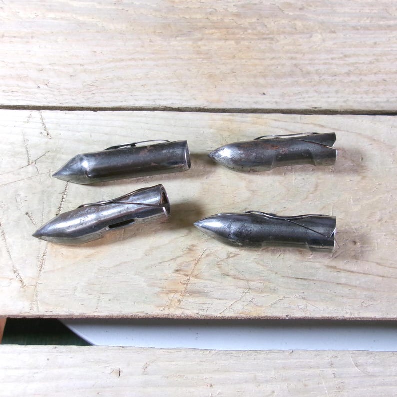 May include: Four vintage, silver-toned metal objects with a pointed, bullet-like shape. Each has a small, curved wire detail. The objects are arranged on a light-colored wooden surface, suggesting a collection or set of tools.
