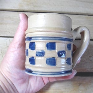 May include: A handmade ceramic mug with a beige base and a blue checkered pattern. The mug has a handle and is held in a hand. The mug is approximately 4 inches tall.