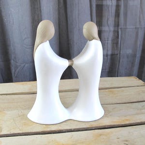 May include: A white ceramic sculpture depicting two figures holding hands. The figures have long, flowing white robes and light brown hair. The sculpture is set against a wooden surface and a gray backdrop.