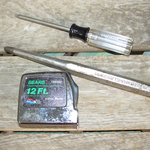 May include: A silver 12-foot tape measure with a green label that reads "SEARS 139382 12 Ft. 1/2-in. Blade".  A silver screwdriver with a black handle and a silver punch tool with the text "USA CRAFTSMAN 84290" are also visible.