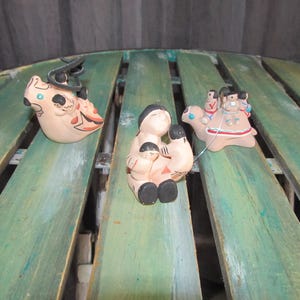May include: Three small ceramic figurines depicting Native American figures. The first figurine is a bird with a person riding on its back. The second figurine is a person holding a child. The third figurine is a turtle with three people riding on its back.