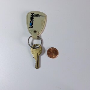 May include: A key with a gold-colored head and a metal ring attached to a cream-colored keychain. The keychain has the word "HANCOR" in blue and the text "BRANDS & WATER MANAGEMENT PRODUCTS". A penny is next to the key.