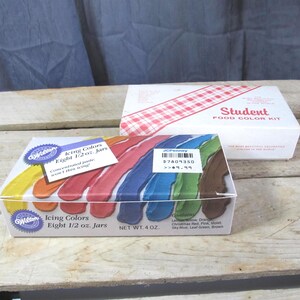 May include: Two boxes of food coloring are displayed on a wooden surface. The front box, labeled "Wilton Icing Colors," contains eight 1/2 oz jars of concentrated paste in various colors. A second box, labeled "Student Food Color Kit," is visible in the background.