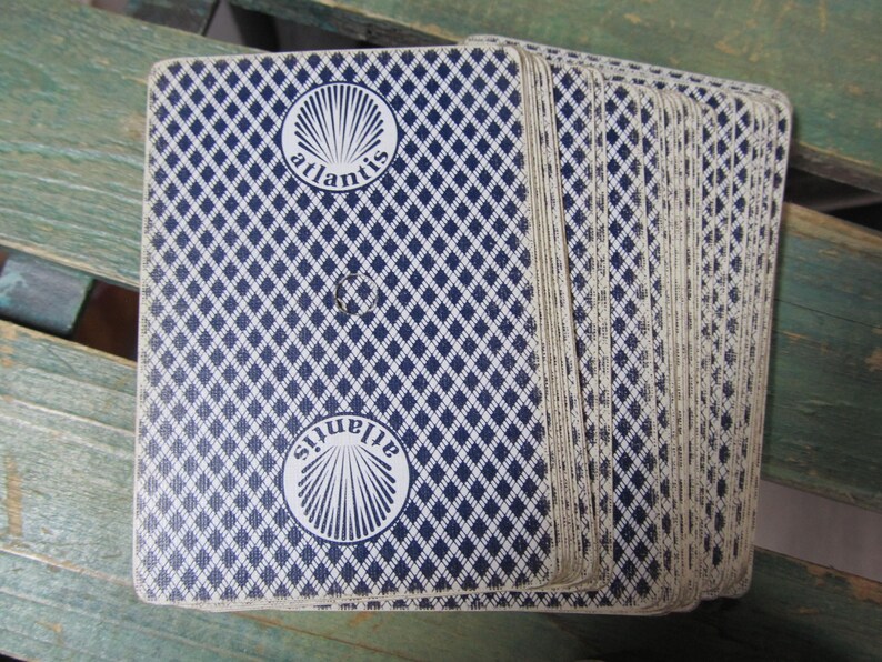 Puede incluir: Una baraja de cartas con un patr&oacute;n de diamantes azules en el reverso. Las cartas tienen la palabra "atlantis" en un c&iacute;rculo blanco. La baraja est&aacute; abanicada, mostrando los bordes de las cartas.