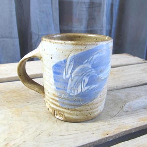 May include: Handmade ceramic mug with a beige and blue design. The mug features a blue fish illustration on a white background. The handle and rim are a speckled brown color. The mug is sitting on a wooden surface.