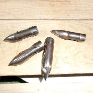 May include: Four antique metal bullet-shaped objects with pointed tips and a silver finish. The objects are arranged on a light brown wooden surface, showcasing their aged appearance and unique design.