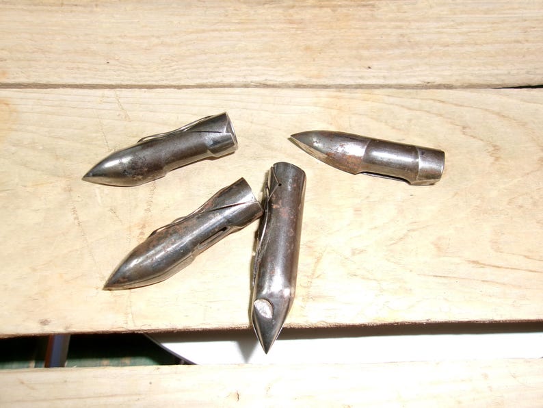 May include: Four antique metal bullet-shaped objects with pointed tips and a silver finish. The objects are arranged on a light brown wooden surface, showcasing their aged appearance and unique design.